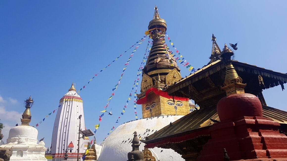 Golden Swayambhunath temple with prayer flags hanging from the top to the bottom with traditional Nepali building in the foreground with angled roof