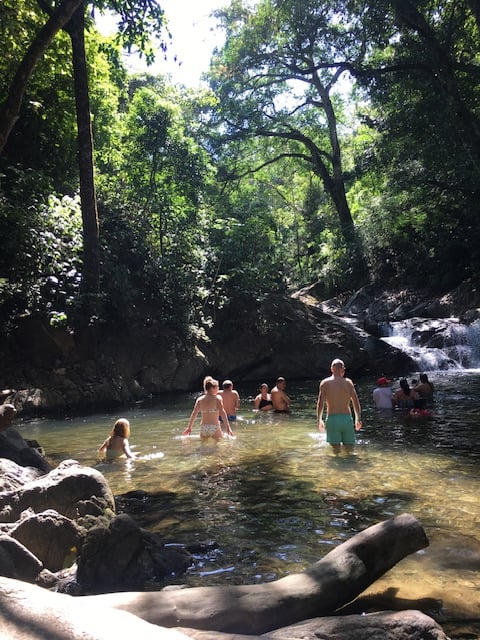 Group of people swimming in a waterfall with the jungle on the back and sun rays lighting up the place.