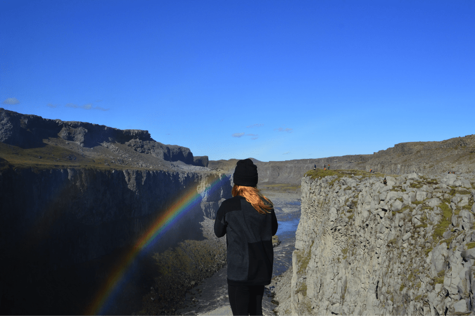 A woman looks out down a massive cliffside where a rainbow can be seen in Iceland