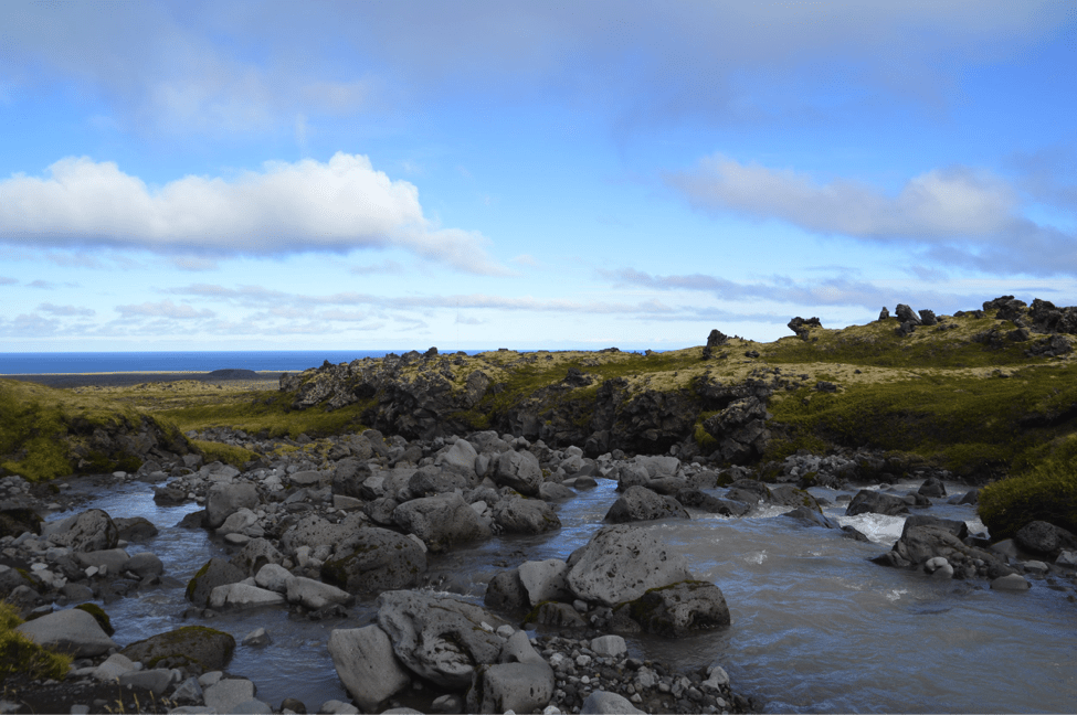 Volcanic rocks lay in a river in Iceland with green hills beyond