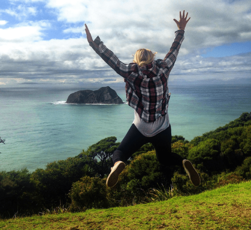 Woman backpacking New Zealand jumps for joy at a pretty ocean view