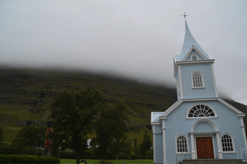 A blue wooden church in Iceland with cloud covered mountains behind
