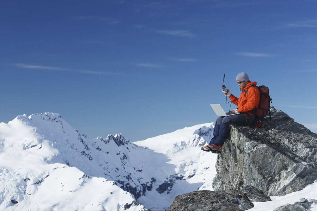 digital nomad in an orange jacket on top of a snowy mountain with a laptop