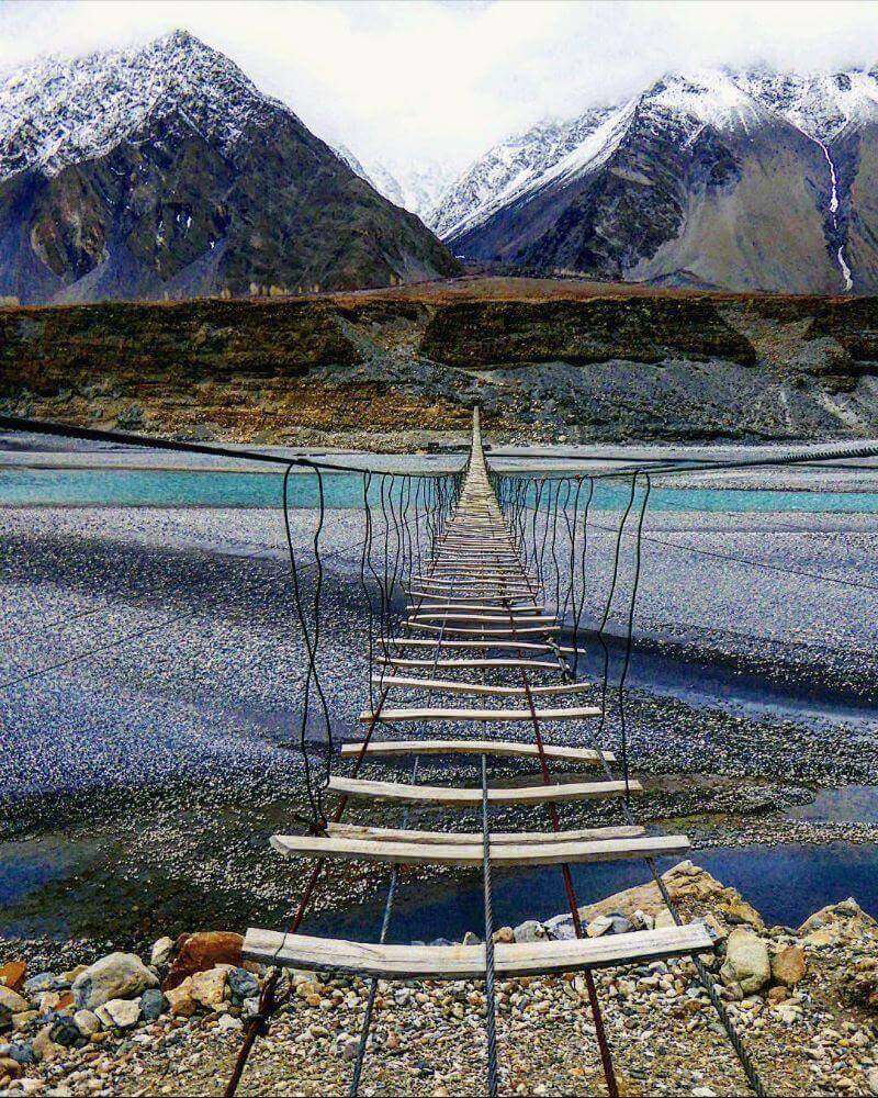 A wooden swing bridge leading out over a river with mountain in the background