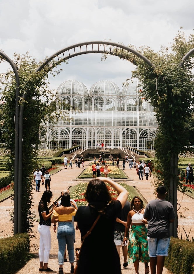 A woman taking a picture  on a botanical garden with people around her and on the background a big construction of a green house.