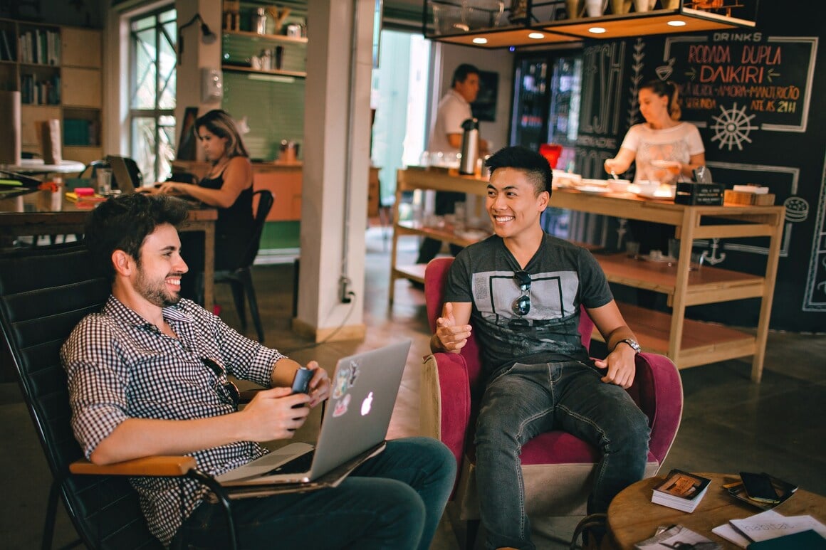 Two boys sitting, one with a computer and both laughing in a hostel. Behind other people working and preparing food.