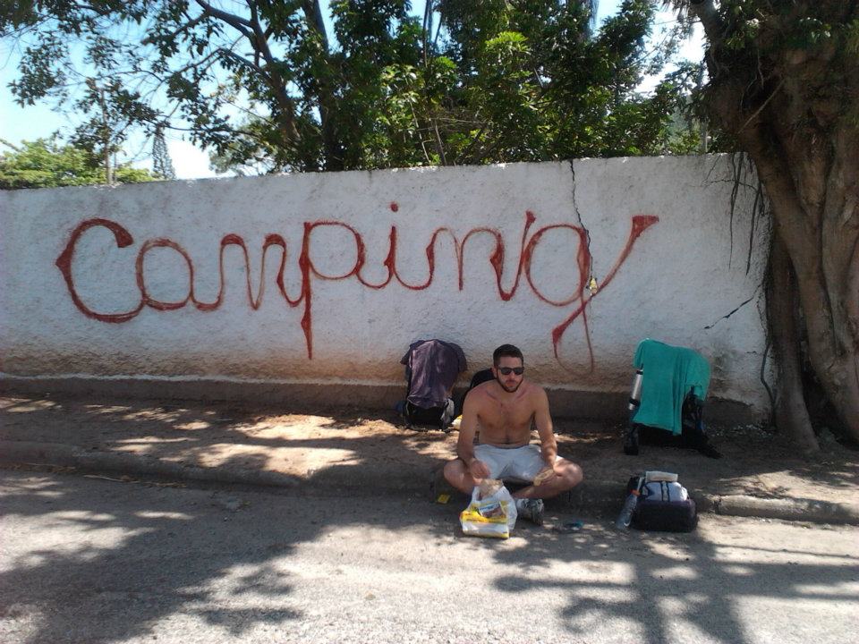 Man waiting topless by the roadside infront of a white wall with 'camping' spray painted in red.