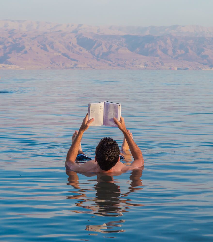 Man floating in the Dea Sea reading a book from the hostel