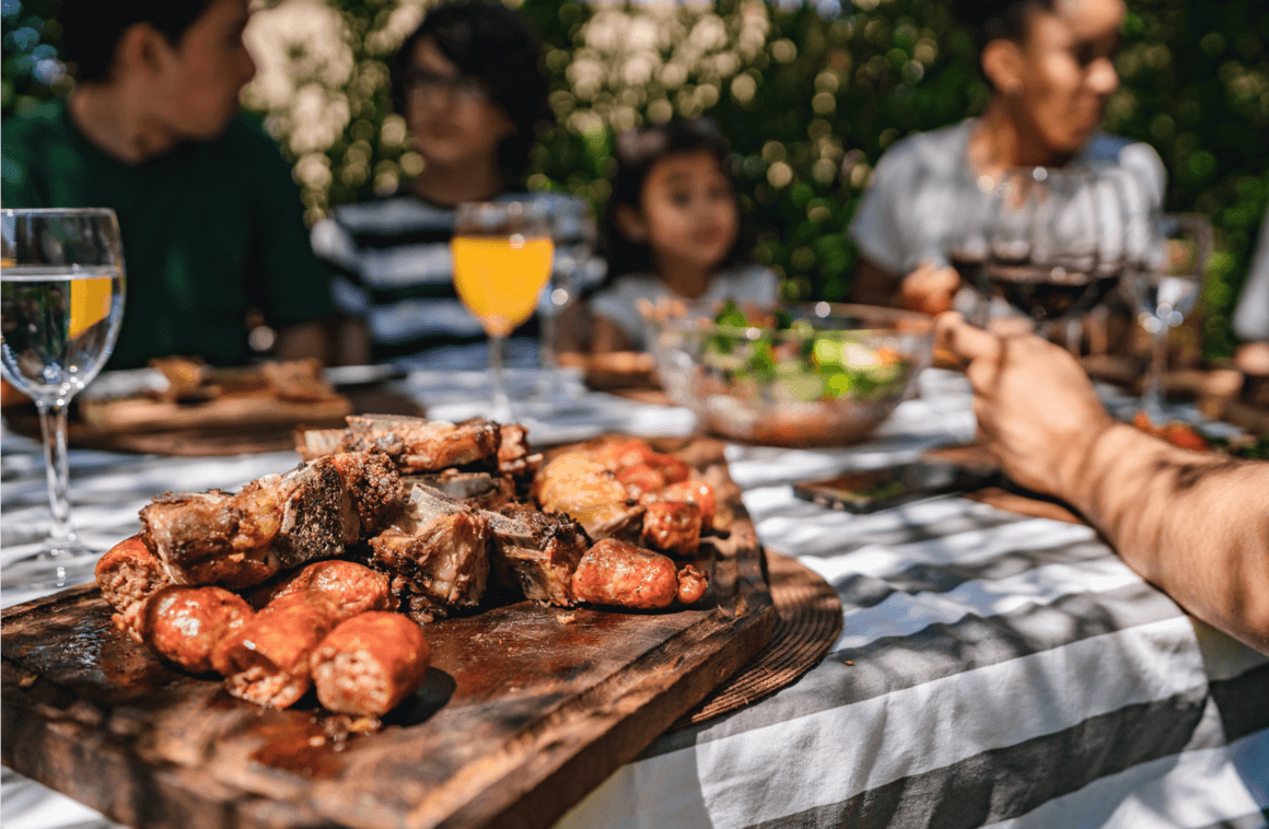 A family sitting around a table and a board of typical Argentine barbecue food in the foreground and some glasses of juice and wine in the background.