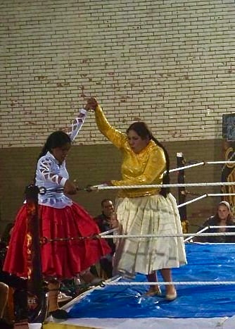 two cholita wrestlers wearing colourful dresses raising their hands together in the ring