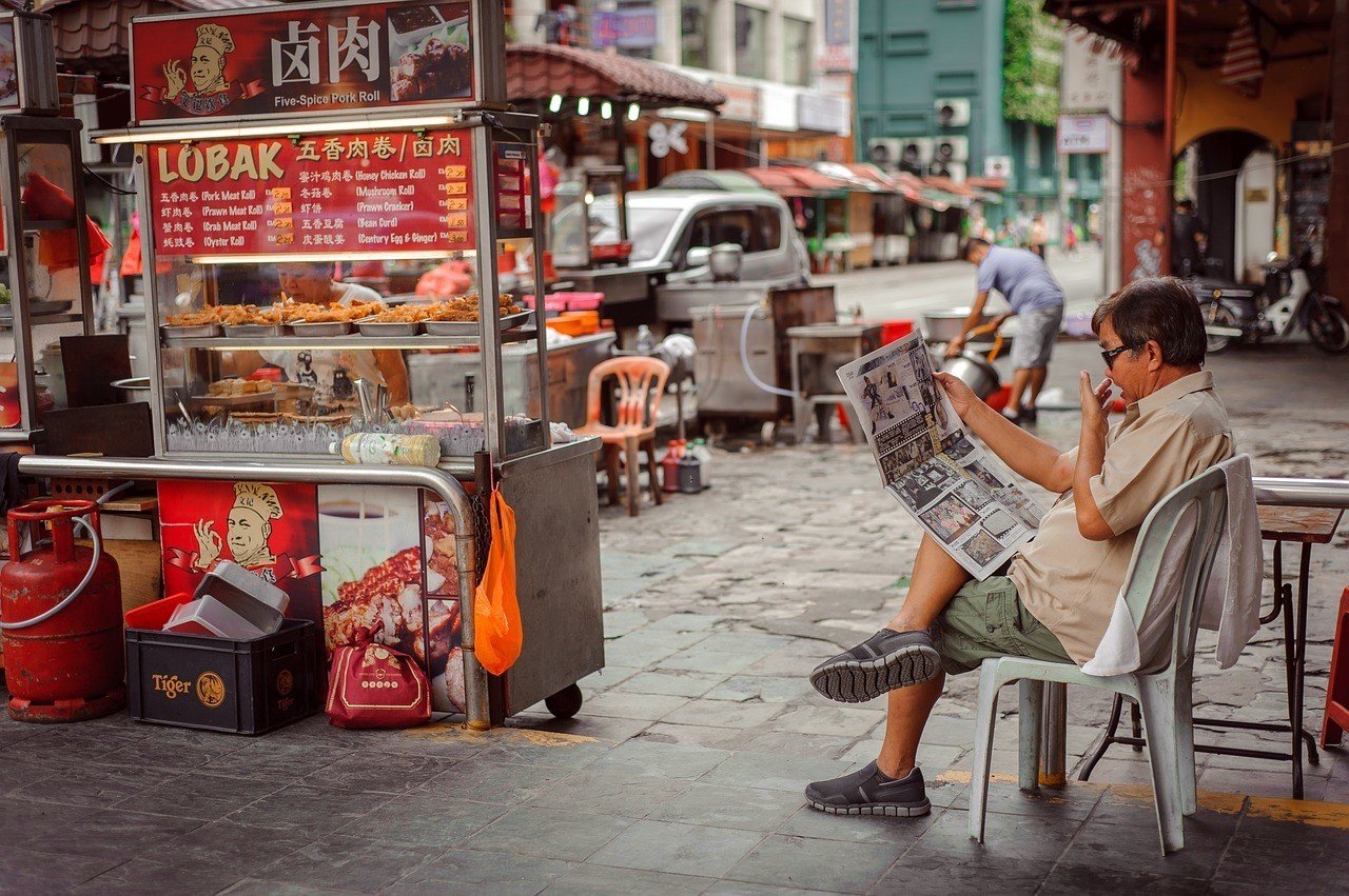 Man on the street reading a newspaper outside a streetfood stand, Chinatown, Kuala Lumpur