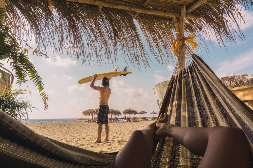 Women laying on a hammock and a man standing on a beach with a surfboard on his head,