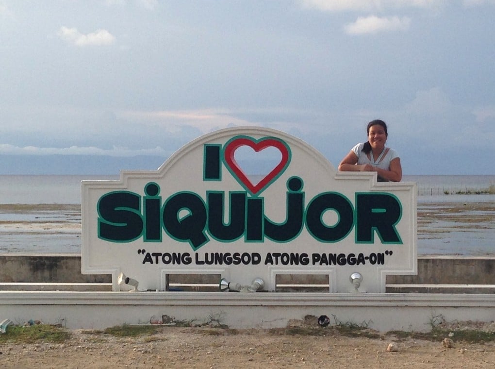 A cheerful girl stands beside a street sign that reads 'I love Siquijor' in the Philippines.