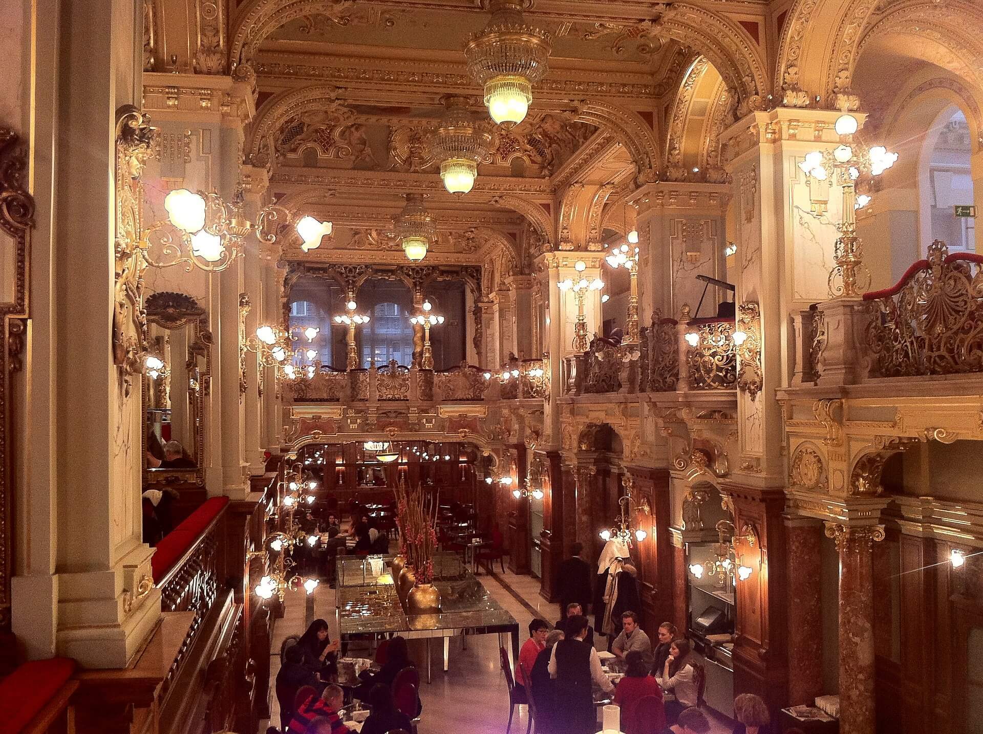 The inside of a fancy, old building with high roofs and chandeliers. People sitting around small tables. 
