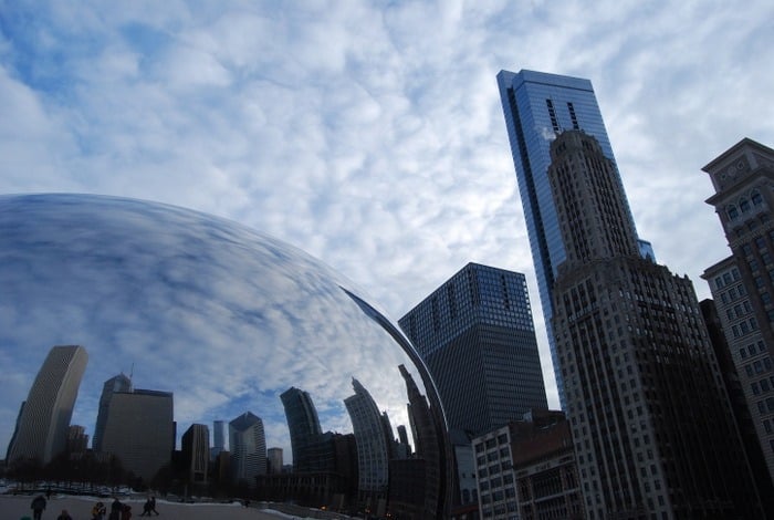 A large, reflective metal sculpture in Millennium Park, Chicago, with the Chicago skyline in the background.