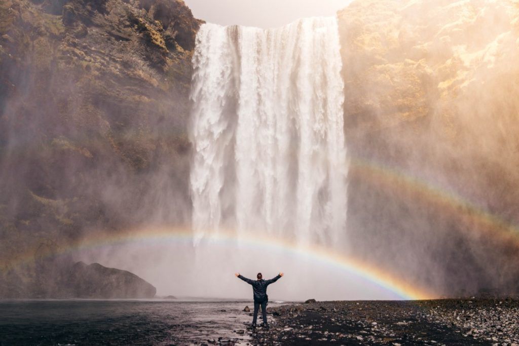 traveller puts hands up in front of waterfall with a rainbow