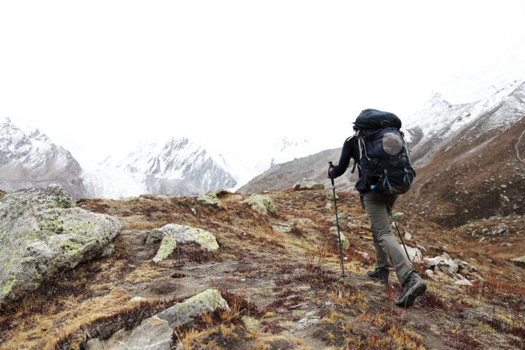 man hiking on a cloudy day on rocky brown terrain toward larege snow covered mountains