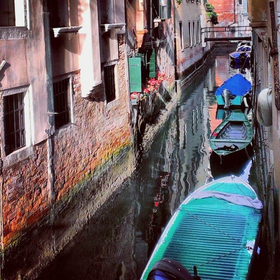 A narrow canal in Venice Italy with several boats lining one wall of the brick and white buildings