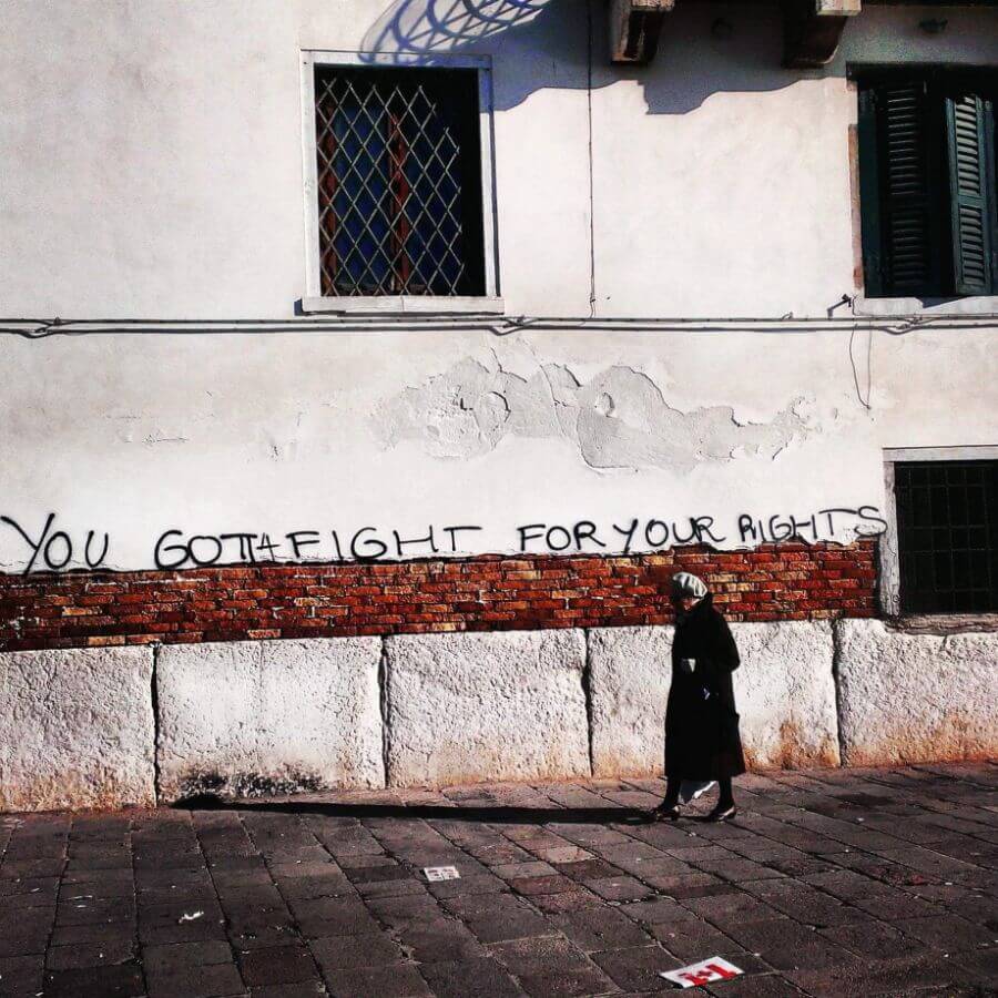A woman walking along a cobblestone street with a white wall that features written street art in Venice Italy