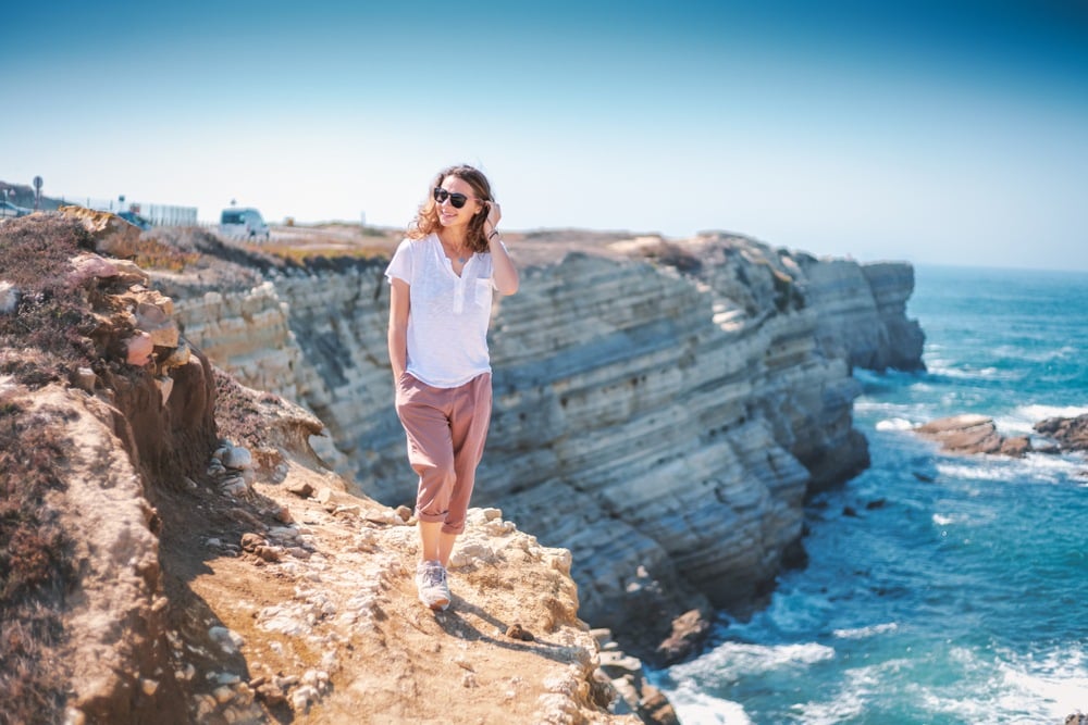 A girl wearing light summer clothes stands on a cliff she is hiking overlooking the sea. 
