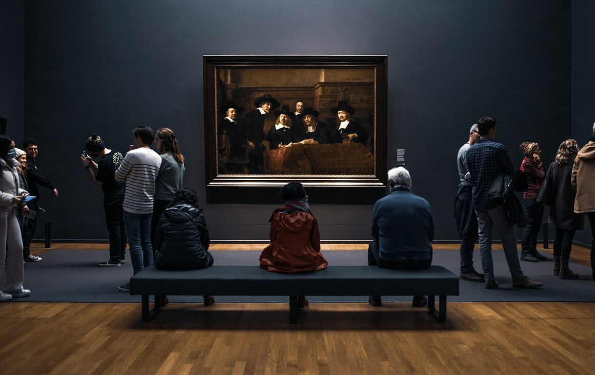People sitting around in the Rembrandt gallery in Amsterdam looking at paintings.