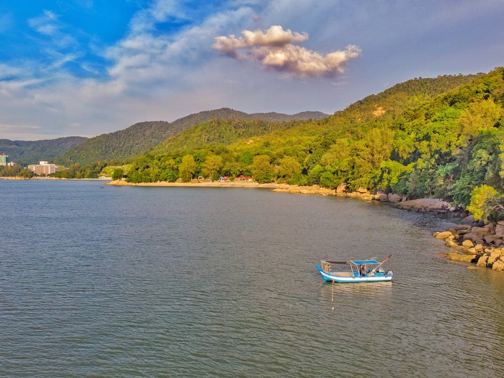 a solo wooden boat on the water in front of forested hills Batu Feringghi Penang