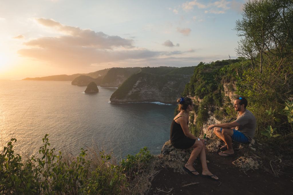 two people sitting on an overlook of the sea in nusa penida at sunset