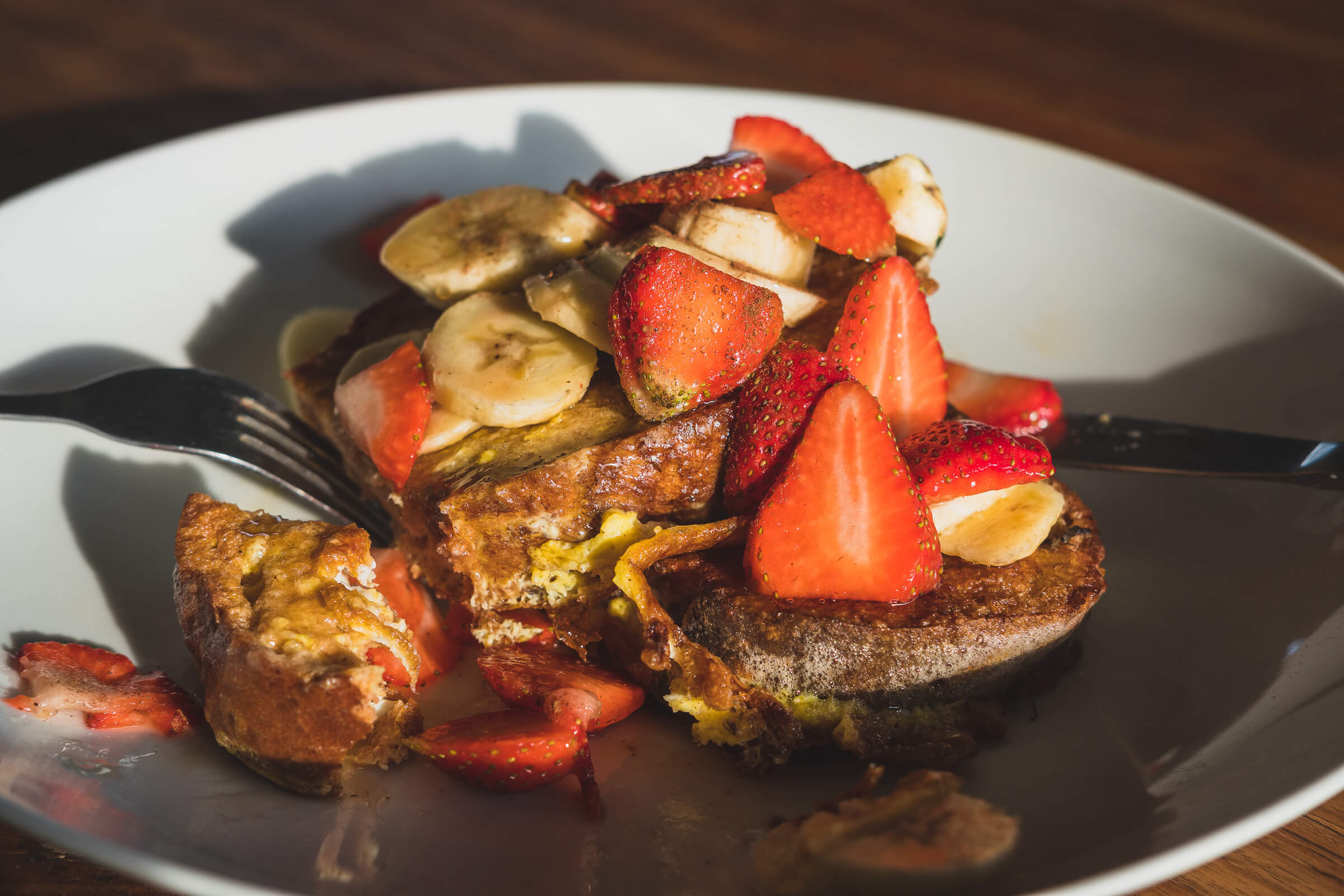 A plate of French toast topped with sliced bananas and strawberries, with a fork on the side and sunlight casting a shadow over the plate