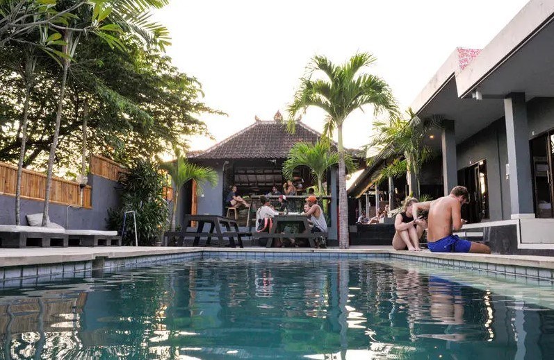 peopkle sitting sround the pool and at the bar at Lay Day Surf best hostels in Canggu with palm trees