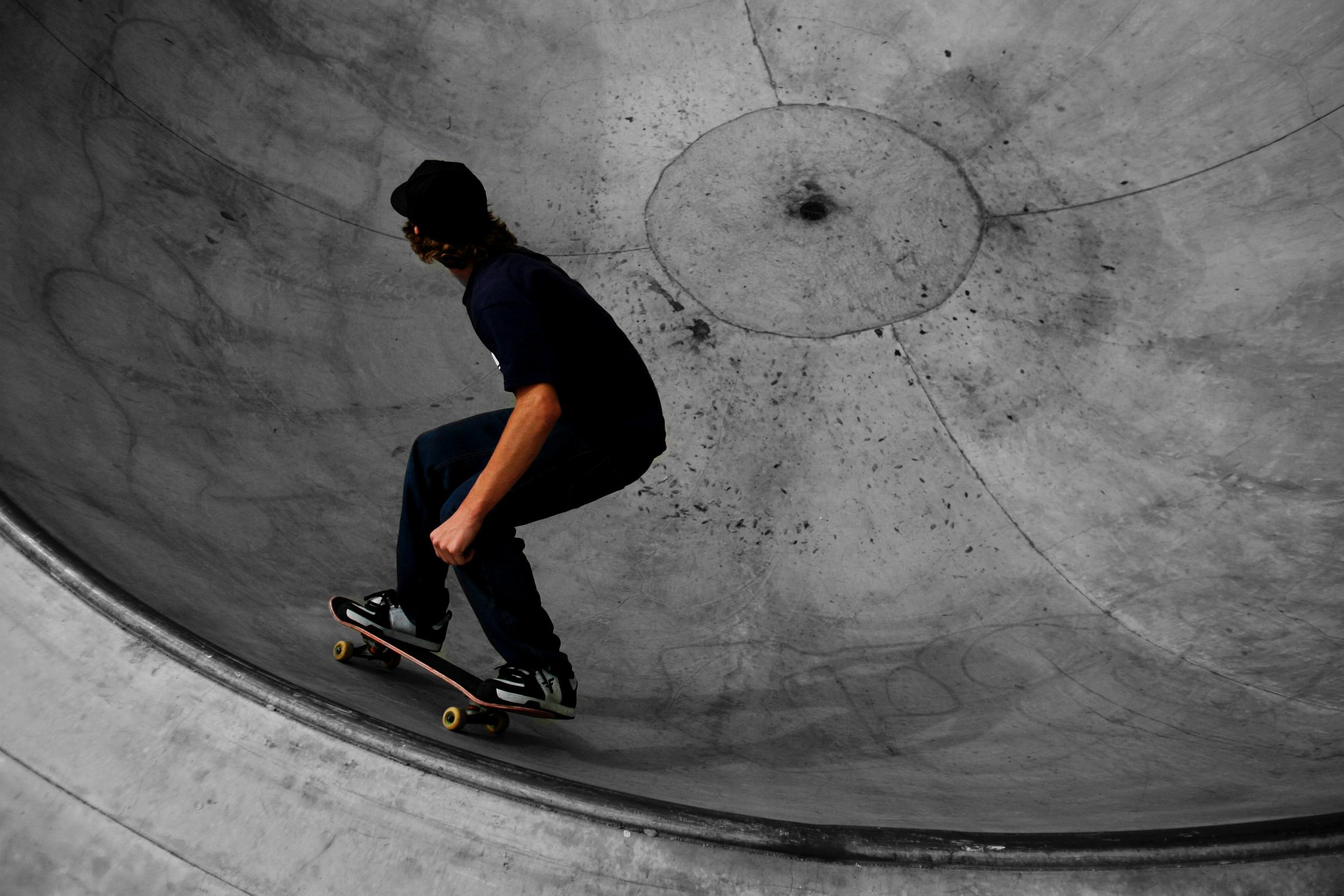A skateboarder riding along the curved interior of a concrete skate bowl. The skateboarder is wearing a dark blue t-shirt and black pants