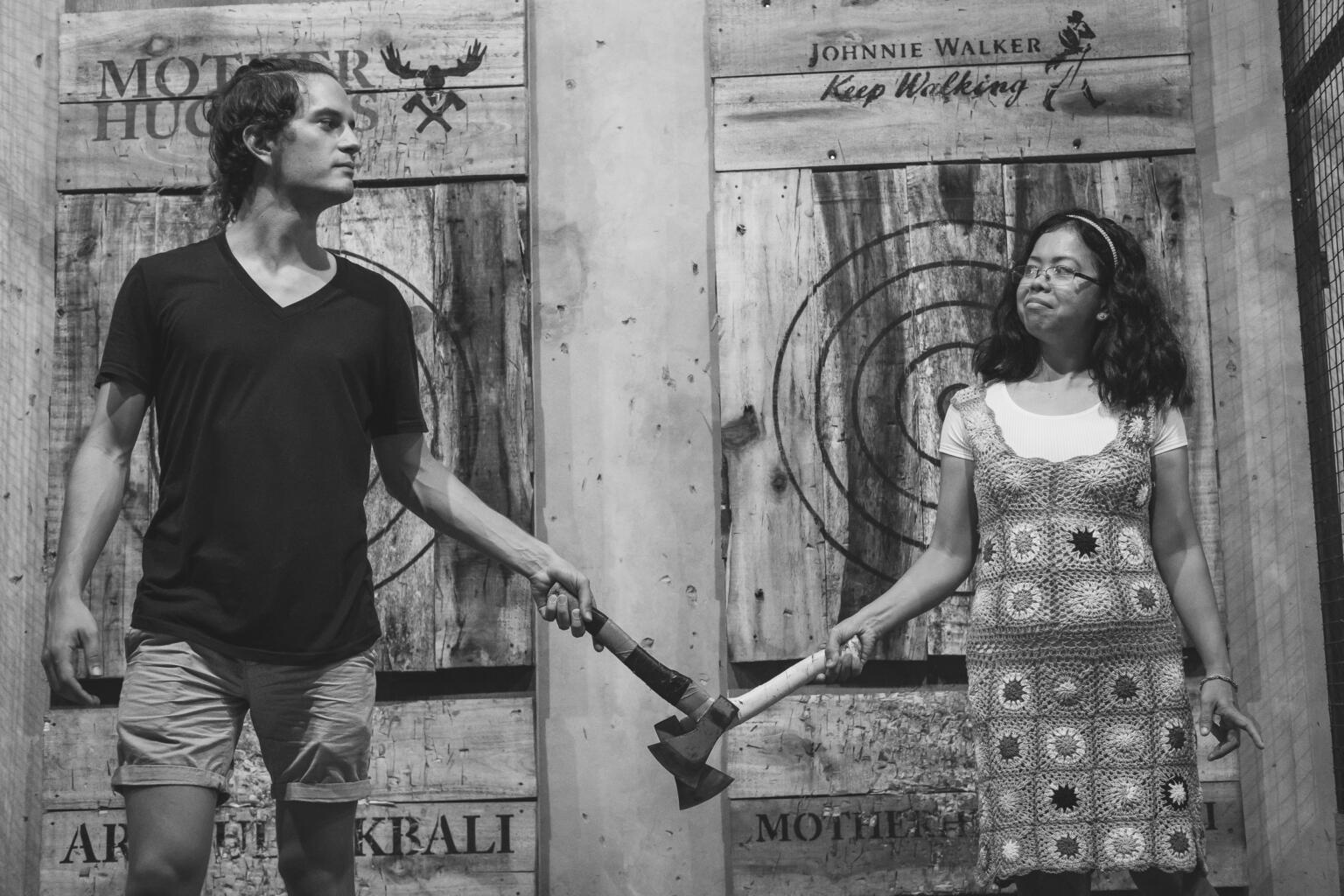 A man and a woman in a black and white photo, both holding an axe together with two hands each. They are standing in front of a wooden background with various logos and a target circle