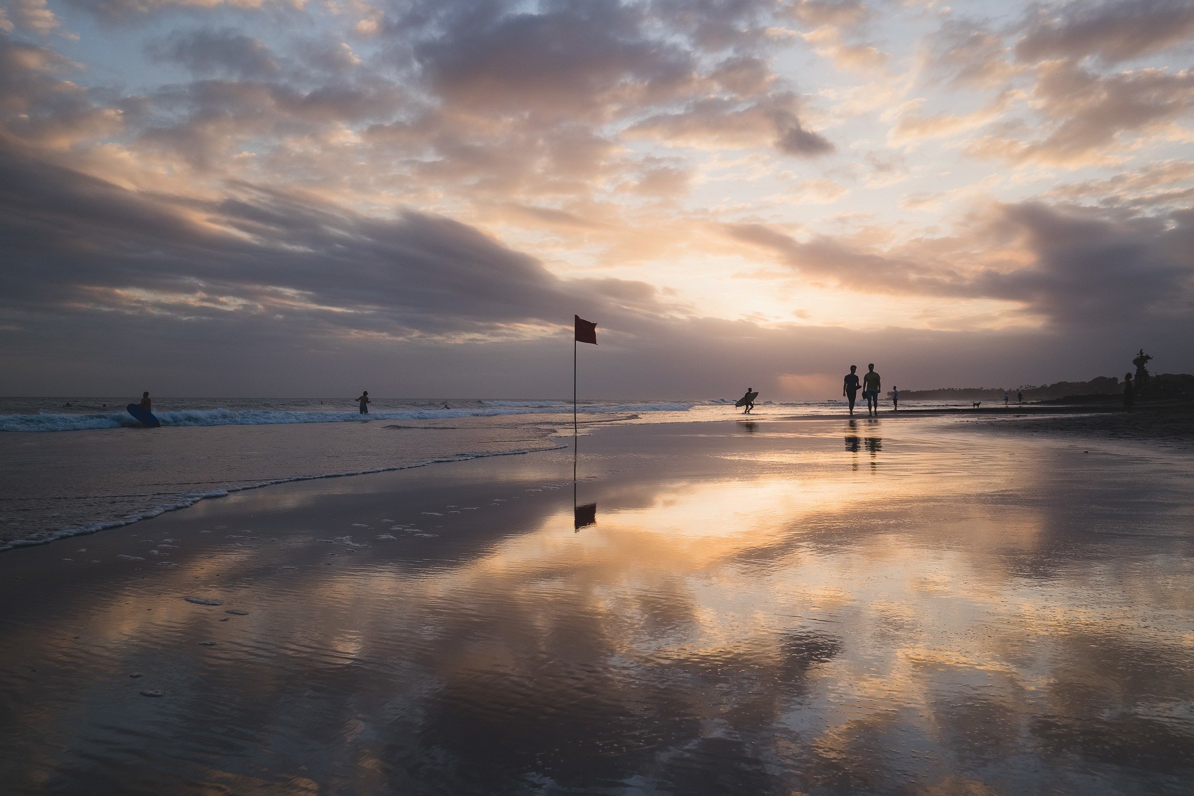 A serene beach scene in Canggu at sunset with a red flag in the foreground, surfers in the water, and silhouettes of people walking along the shoreline, reflecting in the wet sand