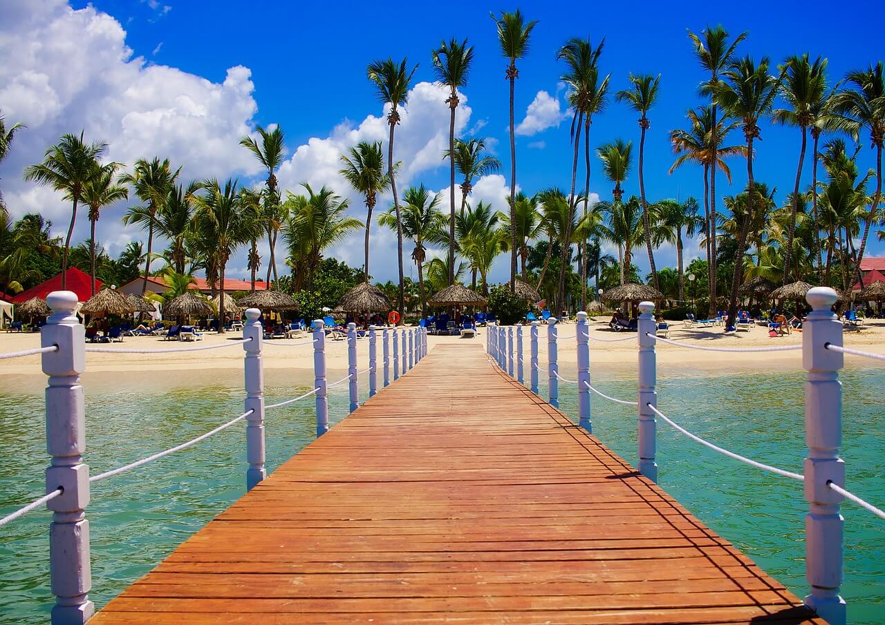 a wooden boardwalk leading to a stretch of sand filled with beach huts and palm trees