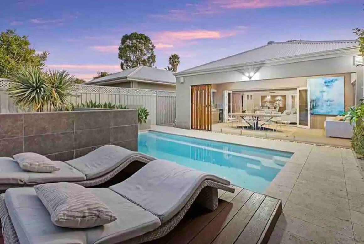 Two pool loungers in the foreground. View of pool and inside the modern house into the living area.