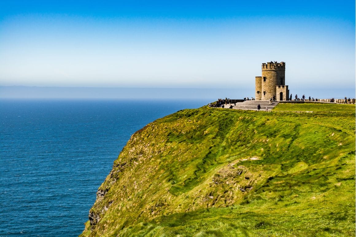 A castle on the edge of a green hill overlooks blue ocean in Ireland. 