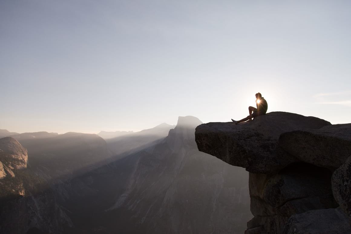 A responsible traveller sits and ponders his life choices on the edge of a cliff at sunrise. 