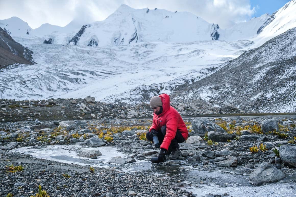 A man in the snow capped mountains fills a waterbottle from a stream