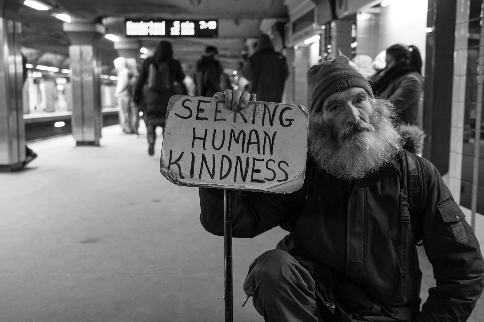 A homeless man begging with a sign saying "SEEKING HUMAN KINDNESS"