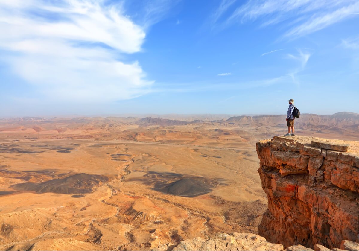 A man stands at the edge of a red cliff overlooking a desert while hiking in the summer. 
