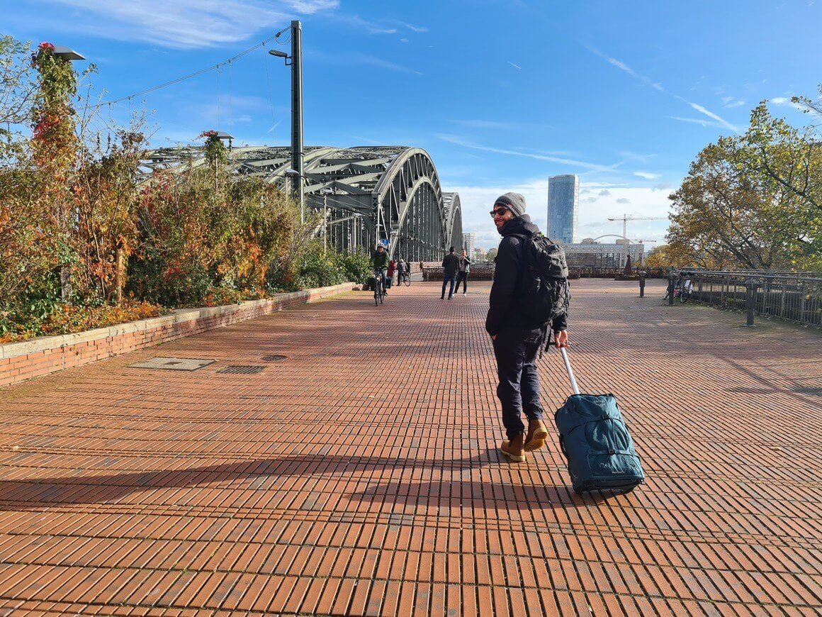 Man walking towards Hohenzollern Bridge in Cologne, Germany in winter clothes wheeling a hand luggage suitcase behind him.