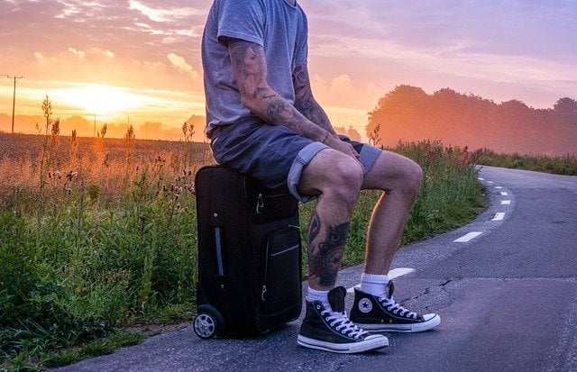 man with travel tattoos sitting on a suitcase by a road with the sunset behind him