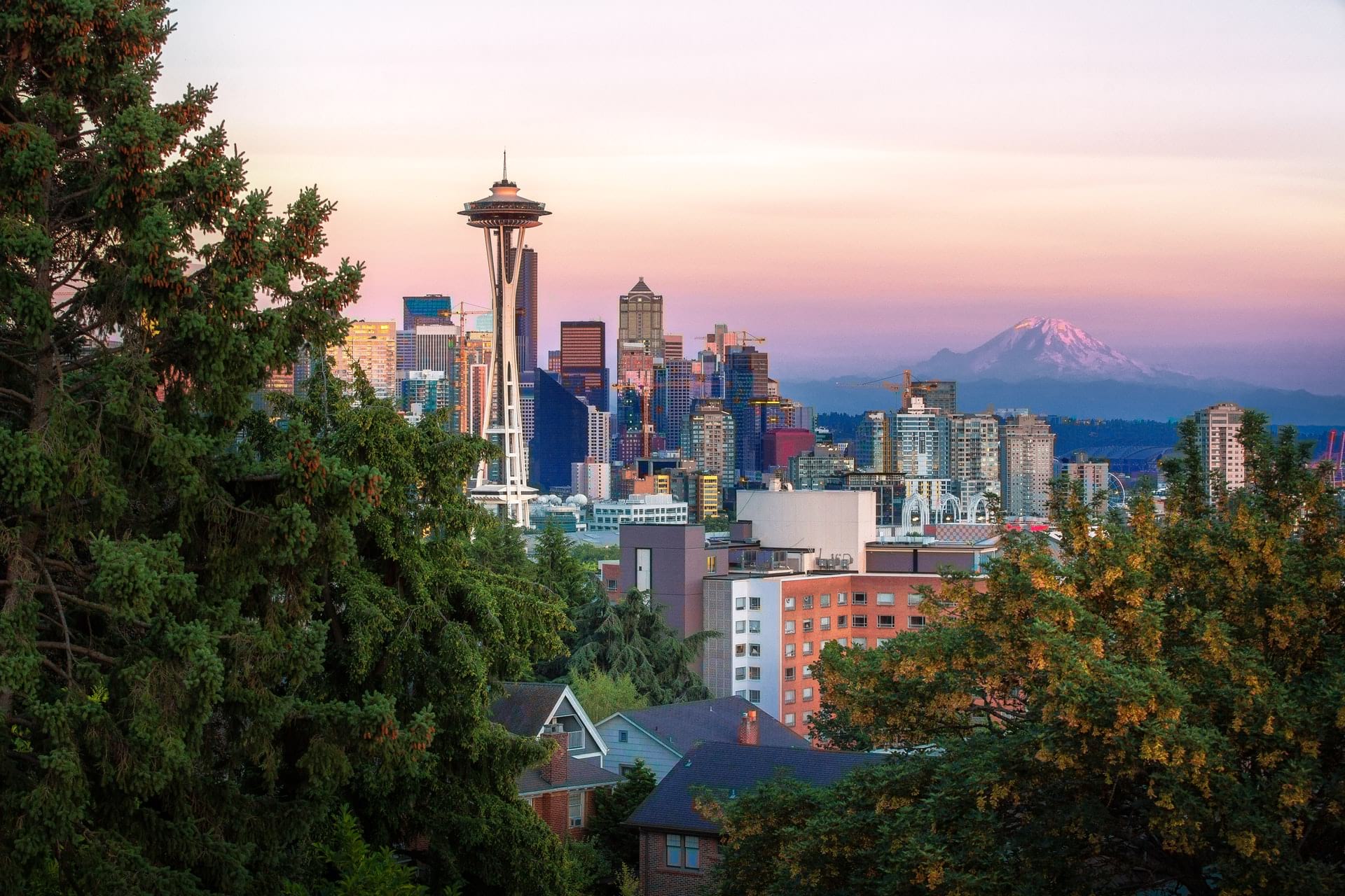 Seattle city skyline, trees in the foreground and mountain peaking up behind the city at sunset