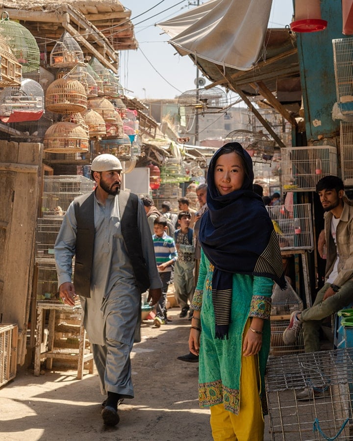 Marsha Jean wears a headscarf in the market of Kabul. There are birds cages and a busy street behind her. 