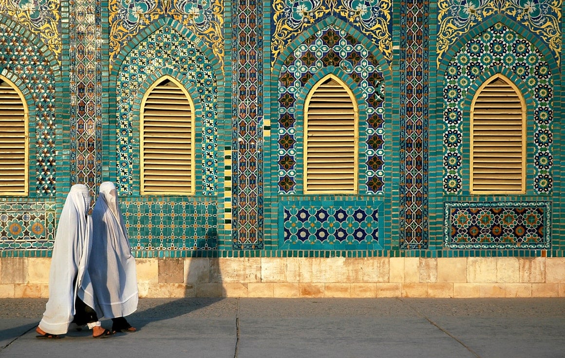 Two women in white hijabs walk past a turquoise tiled mosque. 