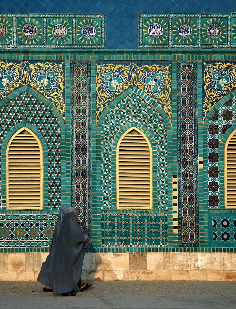 Two women in burqas walk past a turquoise mosque. 