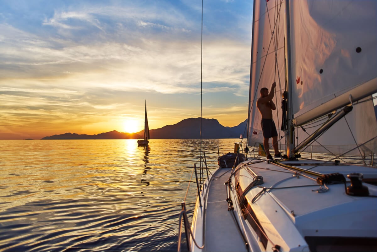 A man stands on his sailboat and raises the mainsail at sunset. 