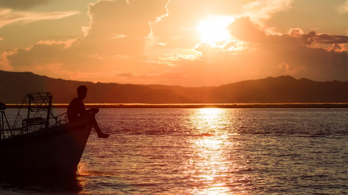 A man sits on the bow of the boat at sunset while crossing the pacific ocean.