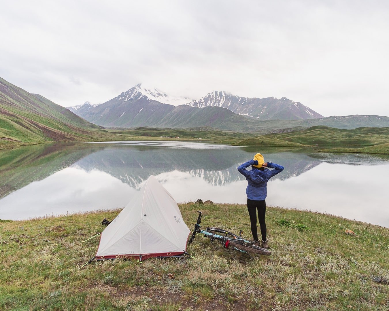 A girl stretches by her tent. Her bicycle is beside her and a picturesque lake and mountains are in front of her. 