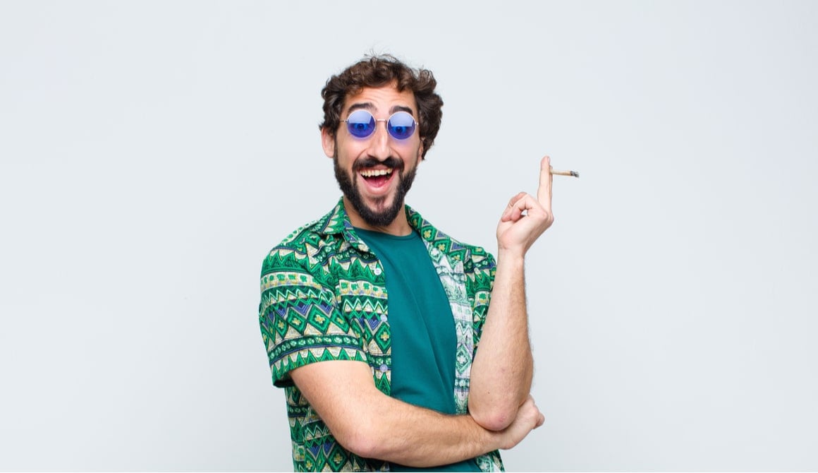 A man with tinted blue glasses holds a joint and smiles goofily. 