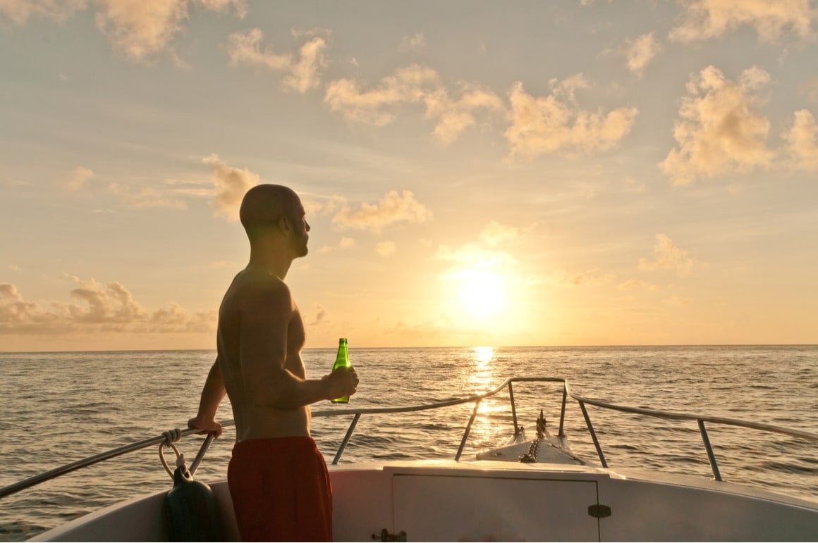 A man stands side on looking at the setting fun with a beer in his hand. 